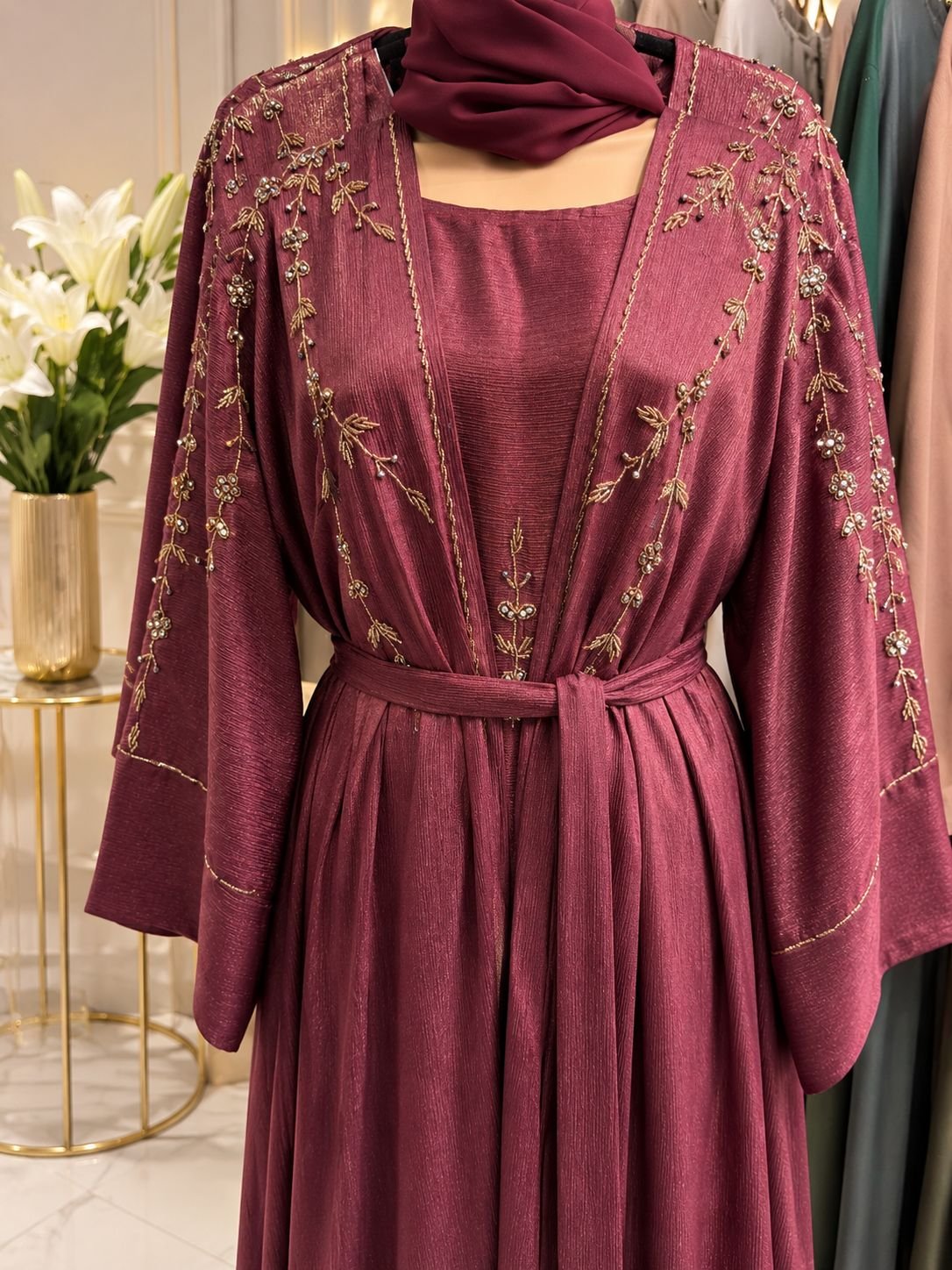 abaya marun coller Elevate your modest wardrobe with this elegant maroon abaya set, crafted from premium textured fabric with a graceful drape. The design features intricate floral embroidery embellished with delicate beadwork along the front panels, shoulders, and sleeves, adding a refined touch of luxury. The open-front abaya is complemented by a matching inner dress and coordinating hijab, creating a sophisticated and effortless look for special occasions, evening gatherings, weddings, Eid celebrations, and formal events. A matching waist belt allows for adjustable styling and a flattering silhouette.